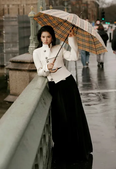 张婧仪撑伞漫步在伦敦雨后街角，白色双排夹克配黑色褶裥半裙，干练摩登的